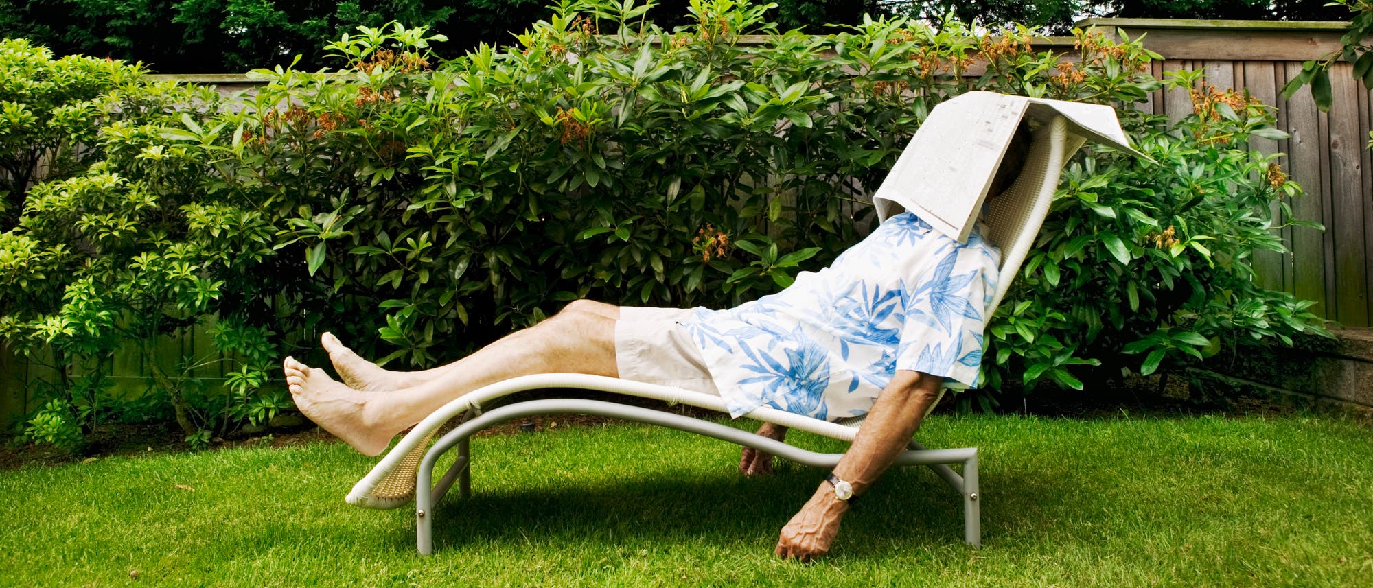 Eine Person liegt entspannt auf einer Gartenliege im Freien. Sie tr&auml;gt ein Hemd mit blauem Blattmuster und helle Shorts. Eine Zeitung bedeckt ihr Gesicht, w&auml;hrend sie sich im Schatten eines Gartens mit &uuml;ppigem Gr&uuml;n ausruht. Im Hintergrund ist ein Holzzaun zu sehen.