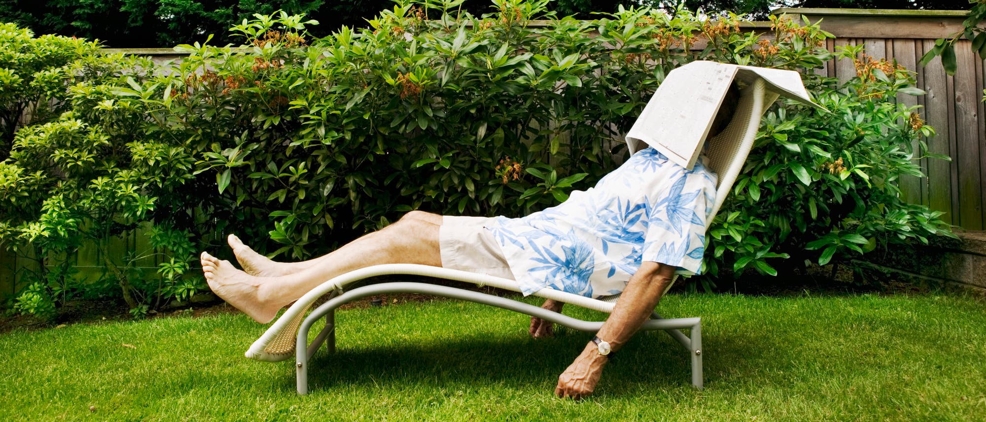 Eine Person liegt entspannt auf einer Gartenliege im Freien. Sie tr&auml;gt ein Hemd mit blauem Blattmuster und helle Shorts. Eine Zeitung bedeckt ihr Gesicht, w&auml;hrend sie sich im Schatten eines Gartens mit &uuml;ppigem Gr&uuml;n ausruht. Im Hintergrund ist ein Holzzaun zu sehen.