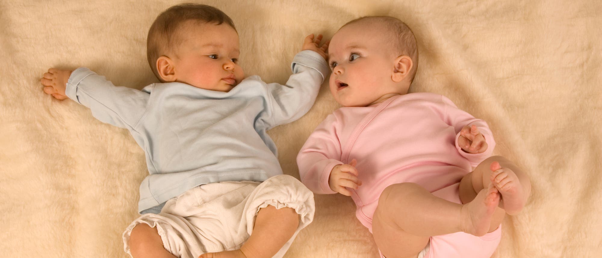 Zwei Babys liegen nebeneinander auf einer weichen, beigen Decke. Das Baby links tr&auml;gt einen blauen Strampler und das Baby rechts einen rosa Strampler. Beide schauen sich an, wobei das Baby rechts den Mund leicht ge&ouml;ffnet hat. Die Szene vermittelt einen ruhigen und neugierigen Moment zwischen den beiden Kindern.