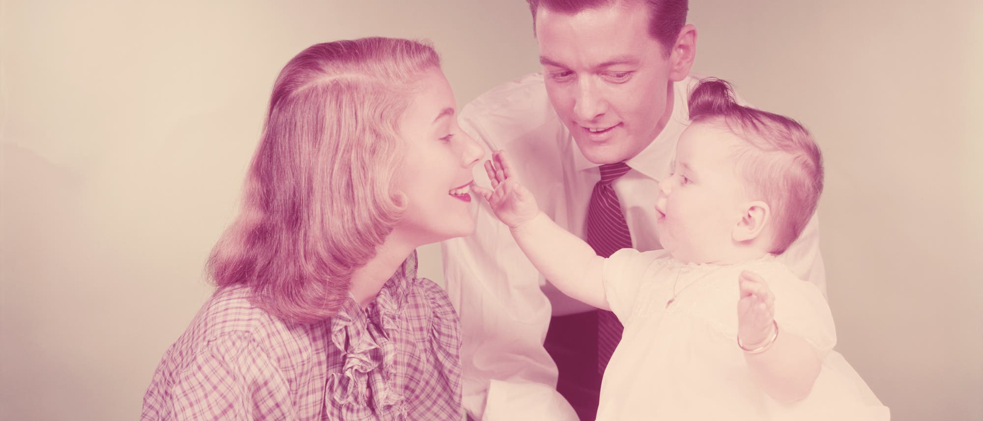 Eine Familie aus den 1950er Jahren in einem Studio-Setting. Ein Baby in einem weißen Kleid sitzt auf einem Tisch und streckt die Hand aus, um das Gesicht der lächelnden Mutter zu berühren. Der Vater in Hemd und Krawatte beugt sich lächelnd zu ihnen. Der Hintergrund ist einfarbig und weich beleuchtet.