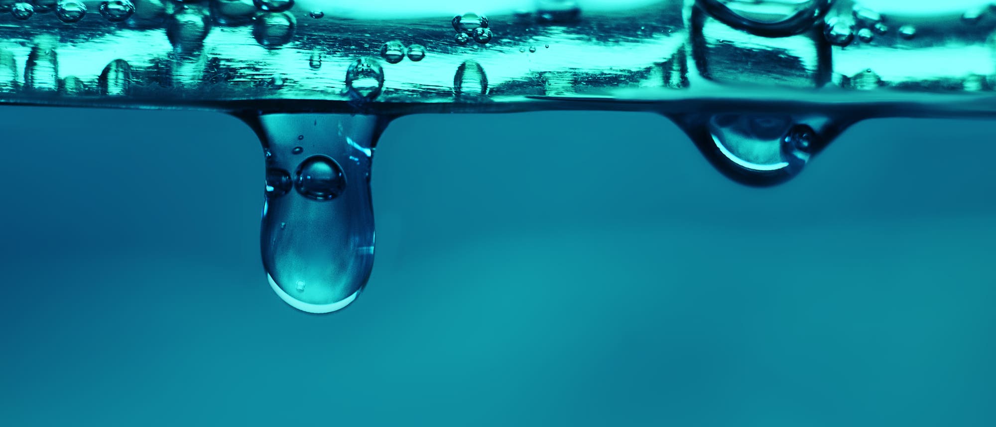 Nahaufnahme eines Wassertropfens, der von einer glatten Oberfl&auml;che herabh&auml;ngt. Der Hintergrund ist in einem sanften Blau gehalten, was die Transparenz und die Lichtbrechung des Tropfens betont. Kleine Blasen sind entlang der Oberfl&auml;che sichtbar, was auf eine fl&uuml;ssige Umgebung hinweist.