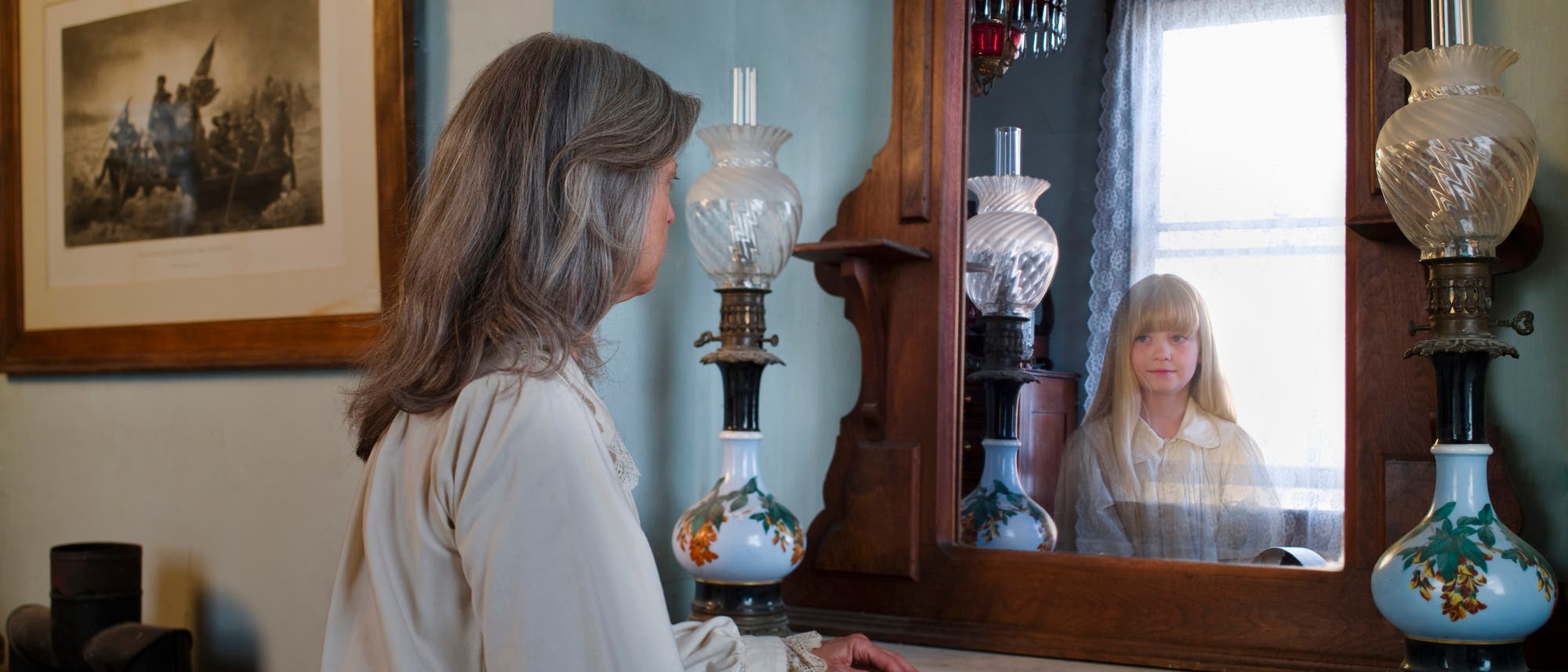 Eine Frau sitzt vor einem antiken Holzspiegel, in dem das Spiegelbild eines M&auml;dchens zu sehen ist. Der Raum ist mit einer alten Lampe und einem gerahmten Bild an der Wand dekoriert. Das M&auml;dchen im Spiegel l&auml;chelt leicht, w&auml;hrend die Frau nachdenklich wirkt. Die Szene vermittelt eine nostalgische und ruhige Atmosph&auml;re.
