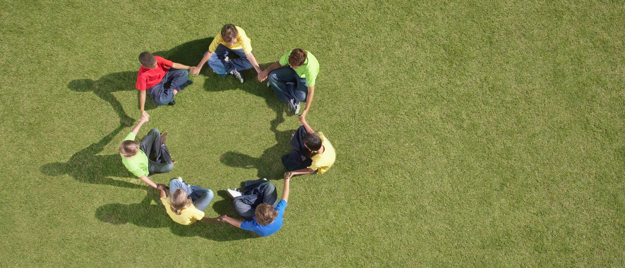 Eine Gruppe von acht Personen sitzt im Kreis auf einer gr&uuml;nen Wiese und h&auml;lt sich an den H&auml;nden. Sie tragen bunte T-Shirts in Rot, Gelb und Gr&uuml;n. Die Szene vermittelt Gemeinschaft und Zusammenarbeit.