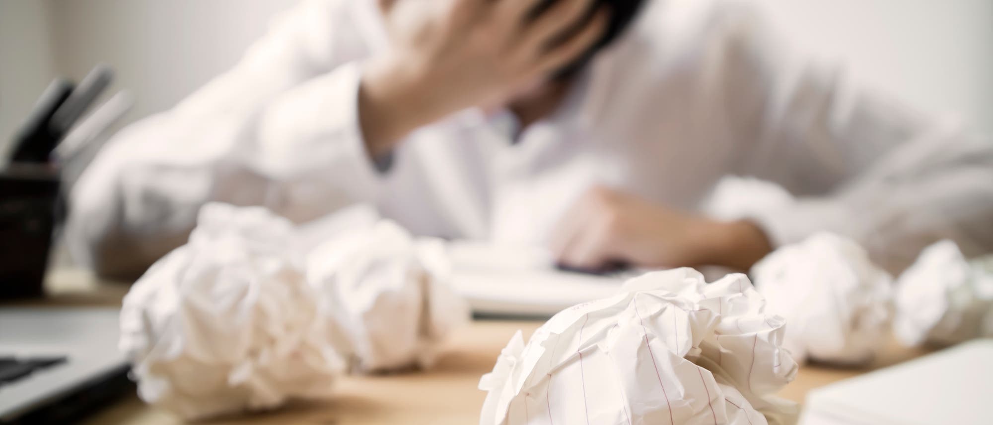 Im Vordergrund zerknüllte Blattseiten, dahinter sitzt ein verzweifelter Mensch vor einem Blatt Papier Im Vordergrund zerknüllte Blattseiten, dahinter sitzt ein verzweifelter Mensch vor einem Blatt Papier