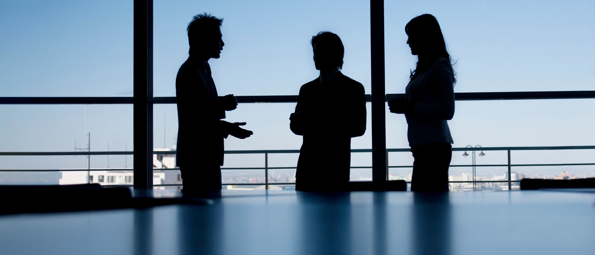 Drei Personen in Business-Kleidung stehen in einem Büro vor einem großen Fenster und unterhalten sich. Die Silhouetten sind vor einem klaren, blauen Himmel sichtbar. Im Hintergrund ist eine Stadtlandschaft zu erkennen. Die Szene vermittelt eine professionelle, geschäftliche Atmosphäre.