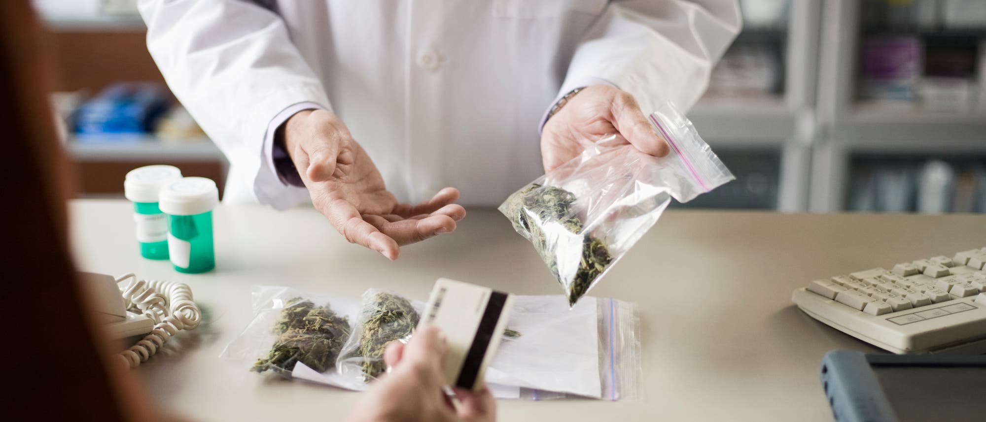 Eine Person in einem weißen Laborkittel übergibt einer anderen Person eine Plastiktüte mit medizinischem Cannabis. Auf dem Tisch liegen weitere Tüten mit ähnlichem Inhalt sowie zwei Pillenflaschen. Die Person im Vordergrund hält eine Kreditkarte. Im Hintergrund sind Regale mit unscharfen Gegenständen zu sehen. Eine Person in einem weißen Laborkittel übergibt einer anderen Person eine Plastiktüte mit medizinischem Cannabis. Auf dem Tisch liegen weitere Tüten mit ähnlichem Inhalt sowie zwei Pillenflaschen. Die Person im Vordergrund hält eine Kreditkarte. Im Hintergrund sind Regale mit unscharfen Gegenständen zu sehen.