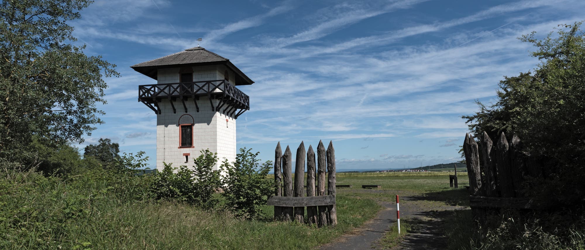 Ein historischer Wachturm aus Stein mit einem Holzdach steht in einer gr&uuml;nen Landschaft. Der Himmel ist blau mit einigen Wolken. Ein schmaler Weg f&uuml;hrt Richtung Turm, am Rand ein kurzes St&uuml;ck eines hohen Zaun aus spitzen Holzpf&auml;hlen, umgeben von Gras und B&uuml;schen.