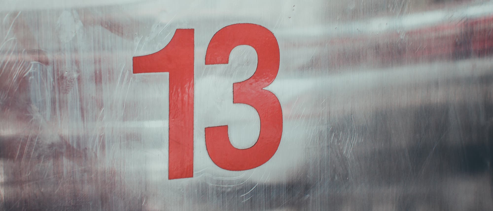 Rote Zahl 13 auf einer metallischen, leicht zerkratzten Oberfl&auml;che. Die Zahl ist gro&szlig; und zentral platziert, was auf eine Kennzeichnung oder Nummerierung hinweisen k&ouml;nnte. Der Hintergrund ist unscharf, was den Fokus auf die Zahl lenkt.