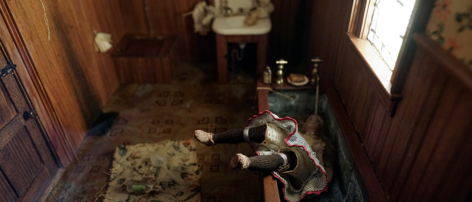 Ein altes Badezimmer mit Holzwänden und einem kleinen Fenster. In der Mitte steht eine Badewanne, in der eine Puppe mit einem rot-weißen Kleid kopfüber liegt, die Beine ragen unnatürlich heraus. Auf dem Boden liegt ein Teppich mit einer Schnapsflasche und einem Glas. Im Hintergrund befindet sich ein Waschbecken mit einigen Gegenständen. Die Szene wirkt düster und altmodisch.
