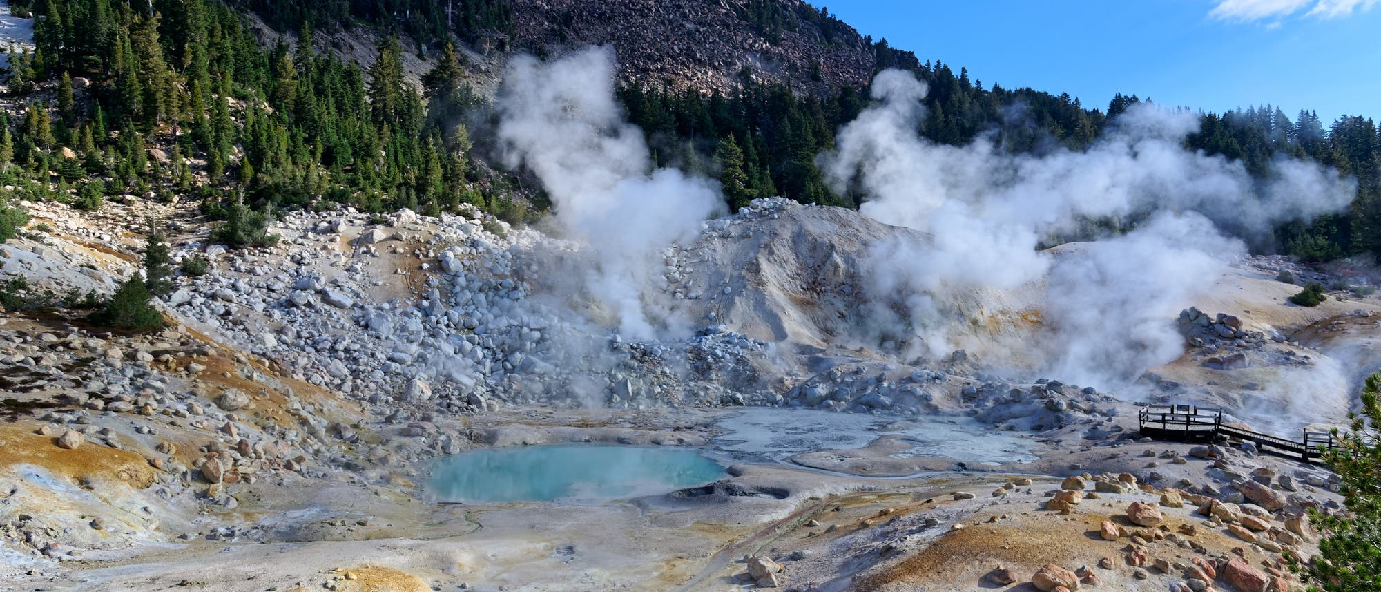 Eine geothermische Landschaft mit dampfenden Fumarolen und einem t&uuml;rkisfarbenen hei&szlig;en Quellbecken, umgeben von felsigem Terrain und Kiefernw&auml;ldern. Im Hintergrund erhebt sich ein bewaldeter H&uuml;gel unter einem klaren blauen Himmel. Eine h&ouml;lzerne Aussichtsplattform bietet einen Blick auf die vulkanische Aktivit&auml;t.