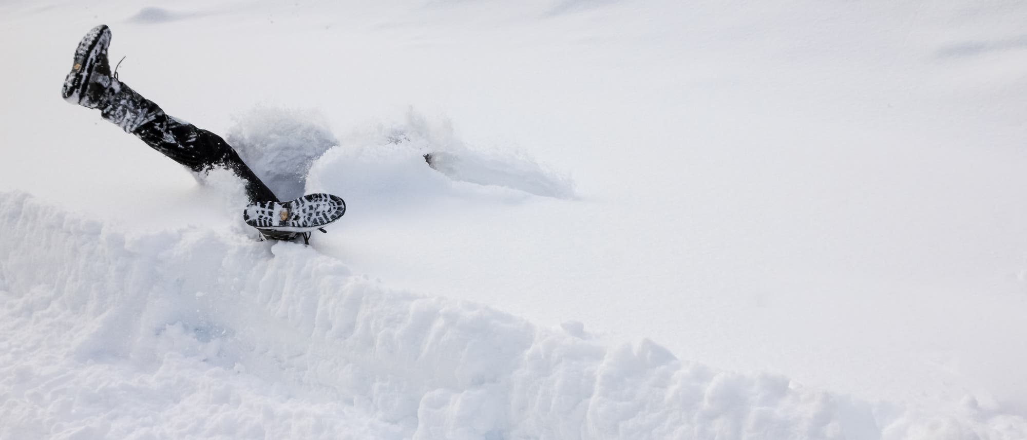 Eine Person ist kopf&uuml;ber in tiefen Schnee gefallen, wobei nur die Beine und Schuhe sichtbar sind. Die Szene zeigt eine humorvolle und unerwartete Situation in einer verschneiten Landschaft. Der Schnee ist unber&uuml;hrt und bildet eine weiche, wei&szlig;e Decke.