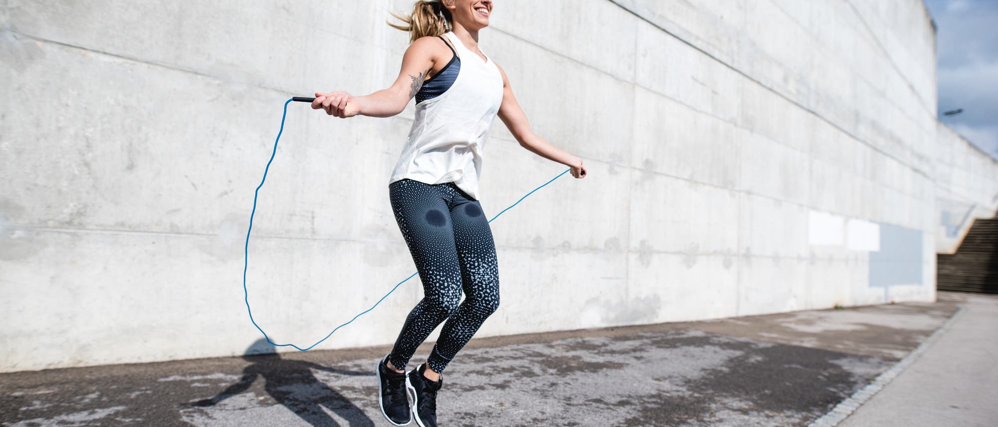 Eine Person springt mit einem blauen Springseil auf einem Bürgersteig vor einer großen Betonwand. Sie trägt ein weißes ärmelloses Oberteil und gemusterte Leggings. Die Sonne scheint, und der Schatten der Person ist auf dem Boden sichtbar. Die Szene vermittelt Energie und Bewegung. Eine Person springt mit einem blauen Springseil auf einem Bürgersteig vor einer großen Betonwand. Sie trägt ein weißes ärmelloses Oberteil und gemusterte Leggings. Die Sonne scheint, und der Schatten der Person ist auf dem Boden sichtbar. Die Szene vermittelt Energie und Bewegung.