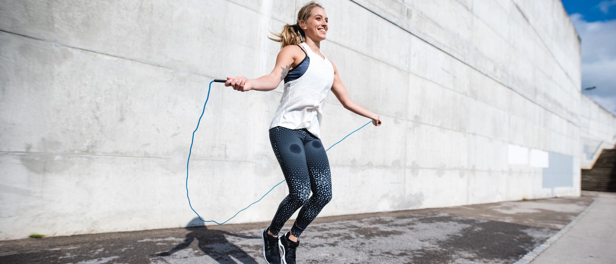 Eine Person springt mit einem blauen Springseil auf einem Bürgersteig vor einer großen Betonwand. Sie trägt ein weißes ärmelloses Oberteil und gemusterte Leggings. Die Sonne scheint, und der Schatten der Person ist auf dem Boden sichtbar. Die Szene vermittelt Energie und Bewegung. Eine Person springt mit einem blauen Springseil auf einem Bürgersteig vor einer großen Betonwand. Sie trägt ein weißes ärmelloses Oberteil und gemusterte Leggings. Die Sonne scheint, und der Schatten der Person ist auf dem Boden sichtbar. Die Szene vermittelt Energie und Bewegung.