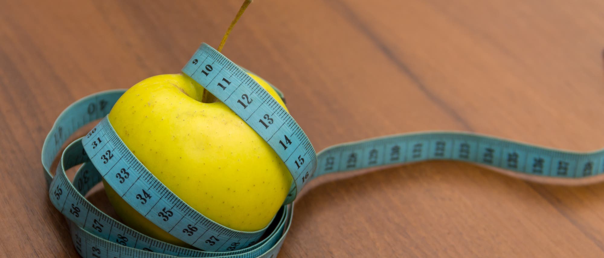 Ein gelber Apfel liegt auf einem Holztisch, umwickelt von einem blauen Maßband. Dieses zeigt Zahlen in Zentimetern.