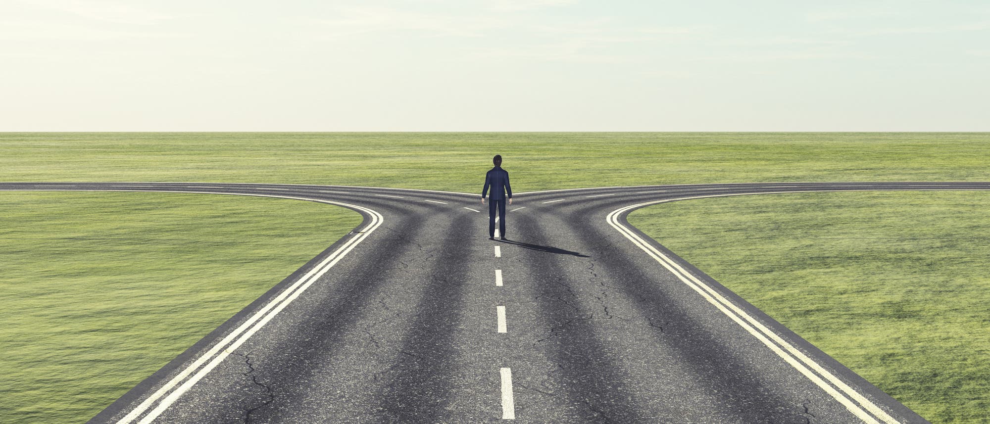 Eine Person steht an einer Weggabelung auf einer asphaltierten Straße, die sich in zwei Richtungen teilt. Die Umgebung ist eine weite, grüne Landschaft unter einem klaren Himmel. Die Szene symbolisiert eine Entscheidungssituation oder einen Scheideweg. Eine Person steht an einer Weggabelung auf einer asphaltierten Straße, die sich in zwei Richtungen teilt. Die Umgebung ist eine weite, grüne Landschaft unter einem klaren Himmel. Die Szene symbolisiert eine Entscheidungssituation oder einen Scheideweg.