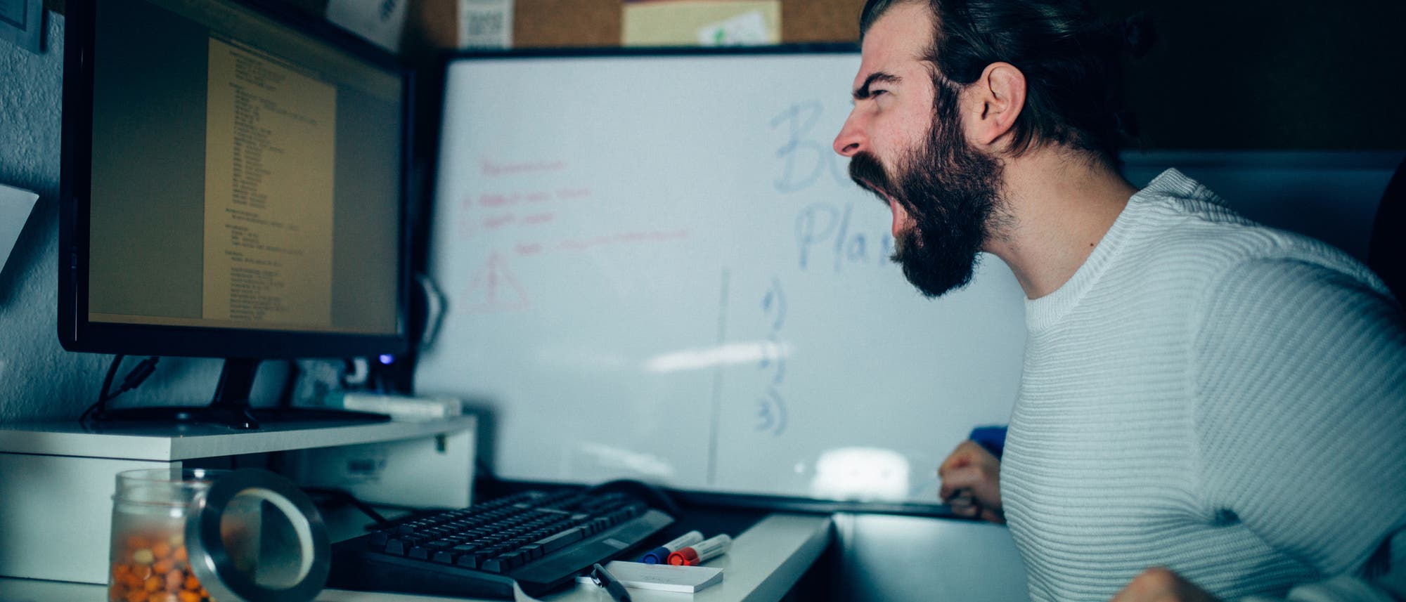 Ein Mann sitzt an einem Schreibtisch vor einem Computerbildschirm und schreit frustriert. Auf dem Bildschirm ist Text zu sehen. Im Hintergrund befindet sich ein Whiteboard mit unleserlichen Notizen. Auf dem Schreibtisch liegen B&uuml;roartikel wie ein Glas mit B&uuml;roklammern und Stifte. Die Szene vermittelt Stress oder Frustration bei der Arbeit.