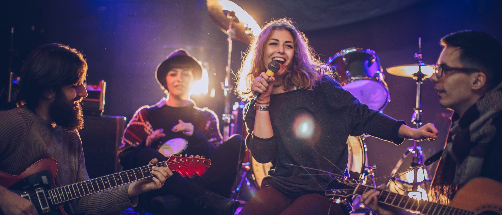Eine Gruppe von vier Musikern probt auf einer Bühne. Eine Frau in der Mitte hält ein Mikrofon und lächelt, während sie singt. Links spielt ein Mann Gitarre, rechts ein weiterer Mann ebenfalls Gitarre. Im Hintergrund sitzt eine Person mit Hut und spielt ein Tamburin. Hinter ihnen sind Schlagzeug und Scheinwerfer zu sehen, die eine lebendige Atmosphäre schaffen. Eine Gruppe von vier Musikern probt auf einer Bühne. Eine Frau in der Mitte hält ein Mikrofon und lächelt, während sie singt. Links spielt ein Mann Gitarre, rechts ein weiterer Mann ebenfalls Gitarre. Im Hintergrund sitzt eine Person mit Hut und spielt ein Tamburin. Hinter ihnen sind Schlagzeug und Scheinwerfer zu sehen, die eine lebendige Atmosphäre schaffen.