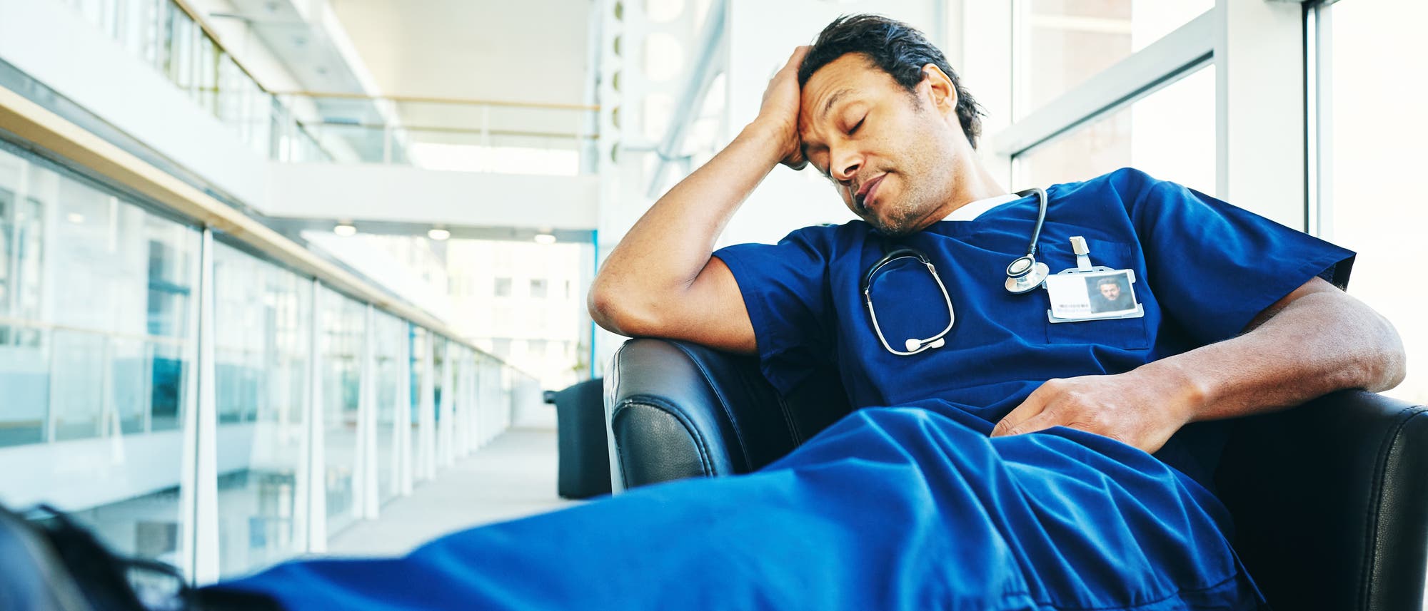 Ein Arzt in blauer Krankenhauskleidung sitzt erschöpft in einem Sessel in einem modernen, lichtdurchfluteten Gebäude. Er hat die Augen geschlossen und stützt seinen Kopf mit einer Hand. Ein Stethoskop hängt um seinen Hals, und ein Namensschild ist an seiner Brust befestigt. Die Szene vermittelt den Eindruck von Müdigkeit und Erschöpfung im medizinischen Beruf. Ein Arzt in blauer Krankenhauskleidung sitzt erschöpft in einem Sessel in einem modernen, lichtdurchfluteten Gebäude. Er hat die Augen geschlossen und stützt seinen Kopf mit einer Hand. Ein Stethoskop hängt um seinen Hals, und ein Namensschild ist an seiner Brust befestigt. Die Szene vermittelt den Eindruck von Müdigkeit und Erschöpfung im medizinischen Beruf.