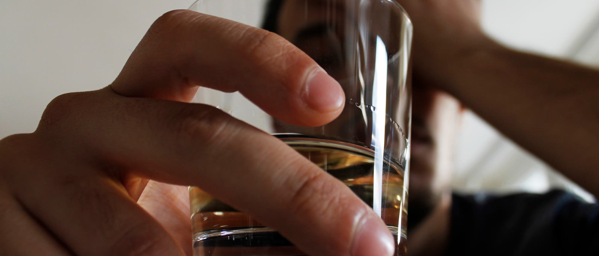 Eine Person h&auml;lt ein Glas mit einer klaren Fl&uuml;ssigkeit, m&ouml;glicherweise Wasser, in der Hand. Die Person st&uuml;tzt ihren Kopf mit der anderen Hand, was auf M&uuml;digkeit oder Stress hindeuten k&ouml;nnte. Der Hintergrund ist unscharf, was den Fokus auf das Glas und die Hand lenkt.