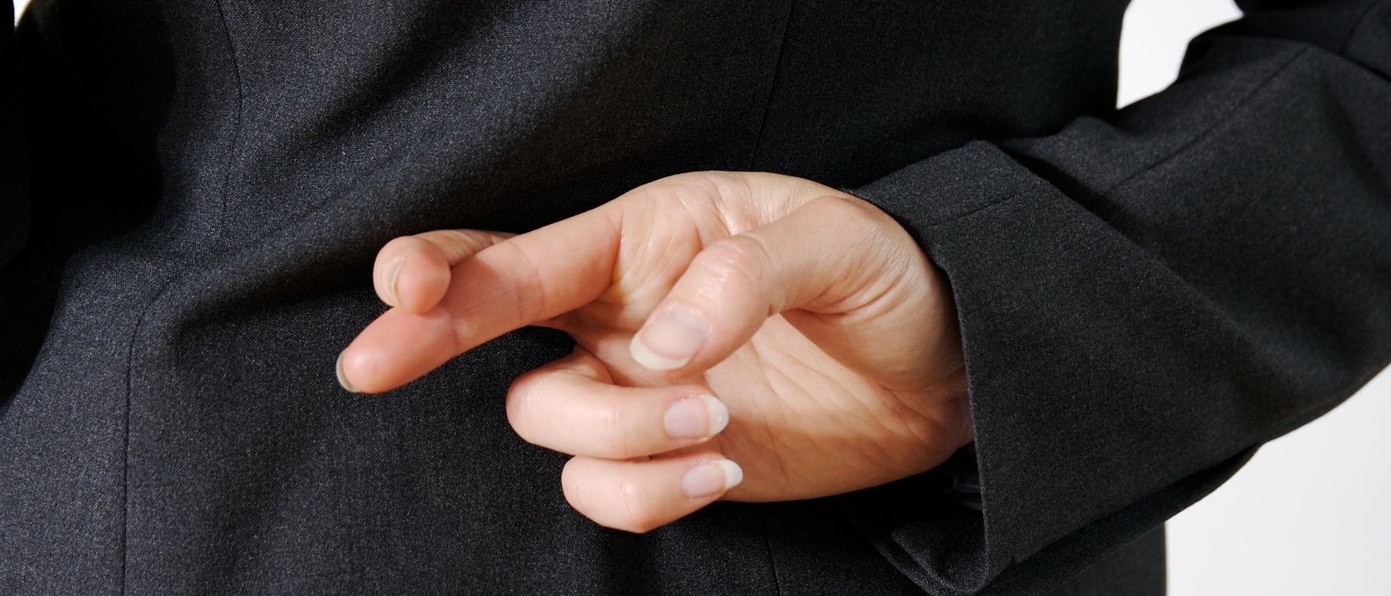 Eine Person in einem dunklen Anzug hält ihre Hand hinter dem Rücken und kreuzt die Finger. Diese Geste wird oft mit Unehrlichkeit assoziiert. Eine Person in einem dunklen Anzug hält ihre Hand hinter dem Rücken und kreuzt die Finger. Diese Geste wird oft mit Unehrlichkeit assoziiert.