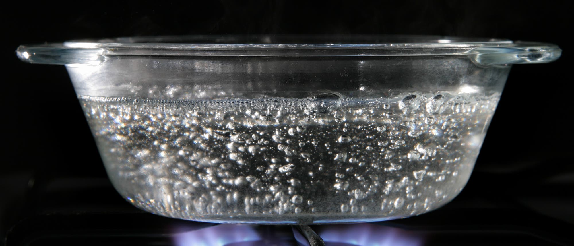 Ein Glasbeh&auml;lter mit kochendem Wasser steht auf einem Gasherd. Der Topf ist mit Blasen gef&uuml;llt, die durch das Erhitzen entstehen. Unter dem Topf ist eine blaue Gasflamme sichtbar, die das Wasser zum Kochen bringt. Der Hintergrund ist dunkel, was den Fokus auf den Topf und die Flamme lenkt.