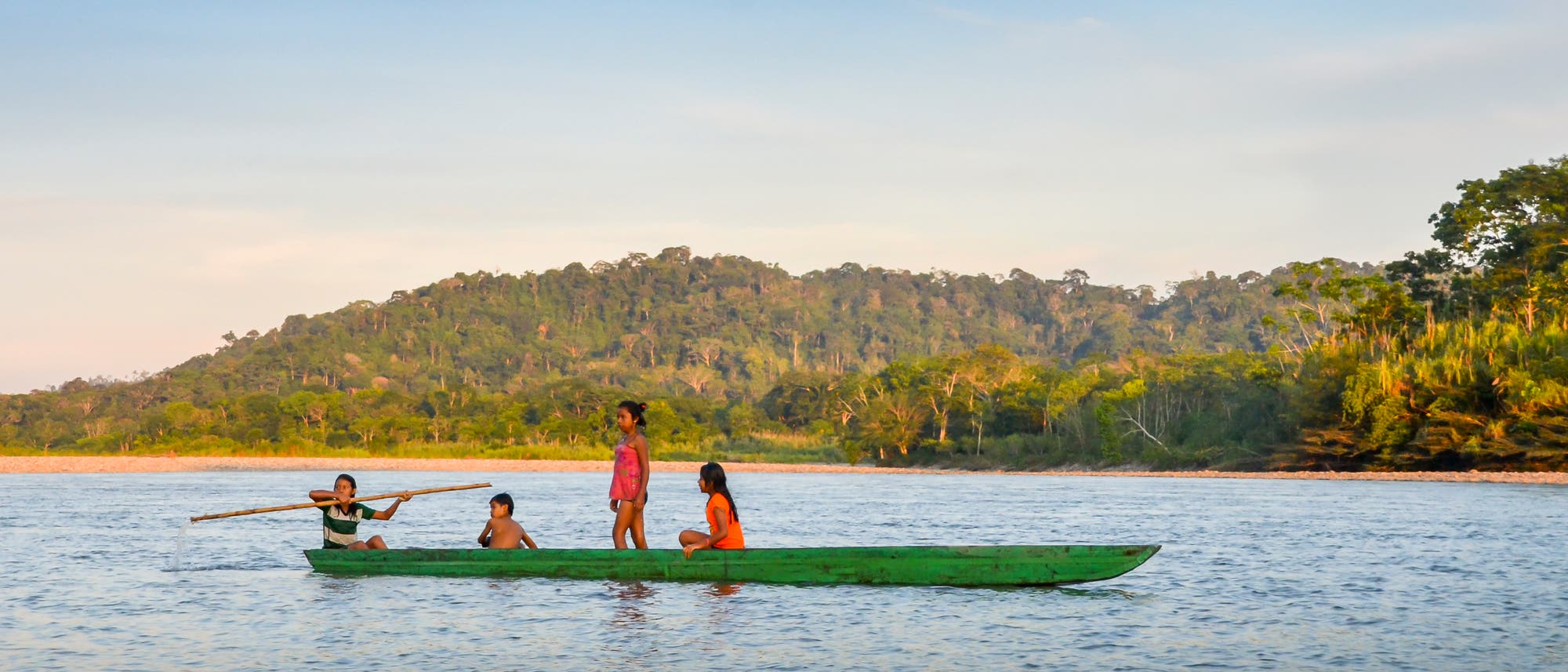 Eine Gruppe von vier Kindern sitzt in einem grünen Kanu auf einem Fluss. Eines der Kinder steht und hält ein Paddel, während die anderen sitzen. Im Hintergrund ist eine bewaldete Hügelkette unter einem klaren Himmel zu sehen. Die Szene vermittelt eine ruhige und natürliche Atmosphäre.