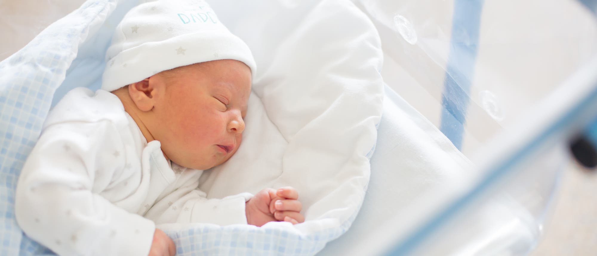 Ein kleines Baby mit Mützchen auf dem Kopf schläft im Bettchen auf der Säuglingsstation.