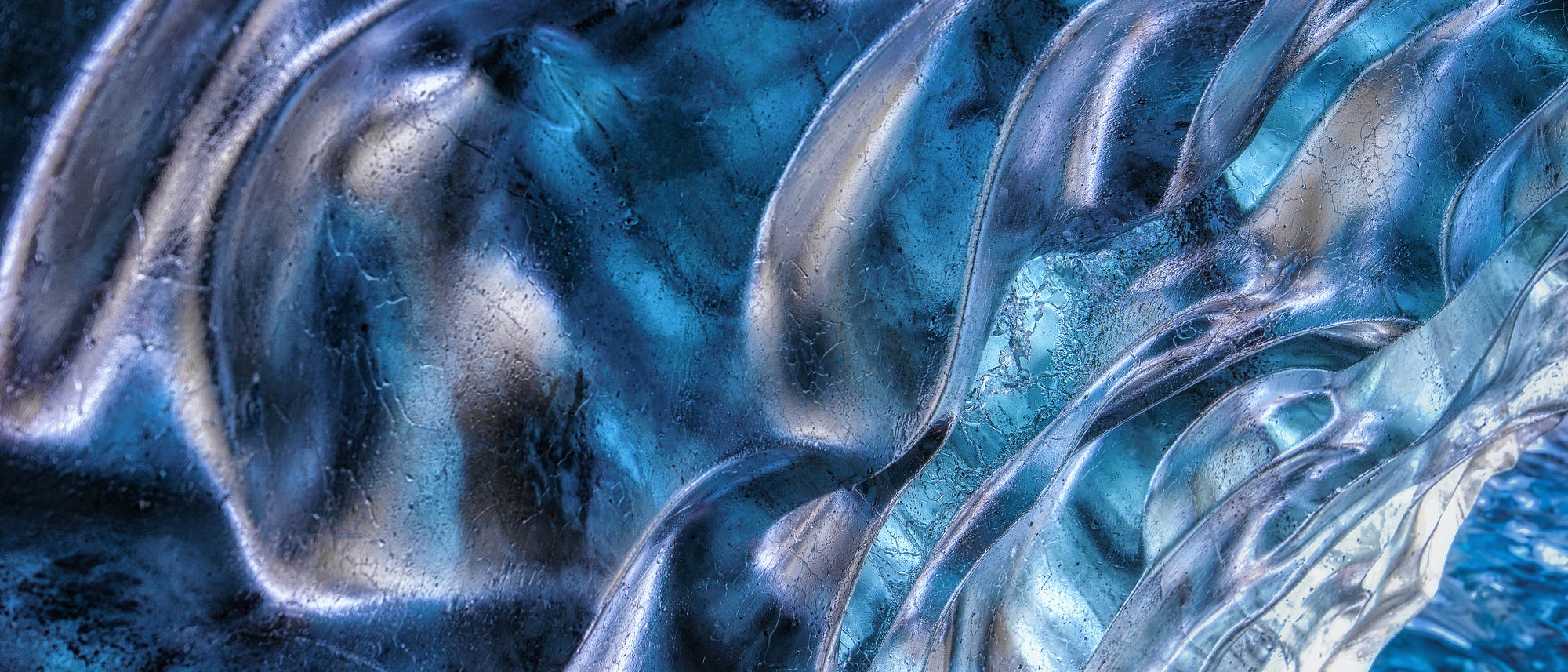 Nahaufnahme von blauen und silbernen Eisstrukturen mit geschwungenen, wellenartigen Mustern. Die Oberfl&auml;che zeigt feine Risse und Texturen, die das Licht reflektieren und eine schimmernde, fast transparente Erscheinung erzeugen.