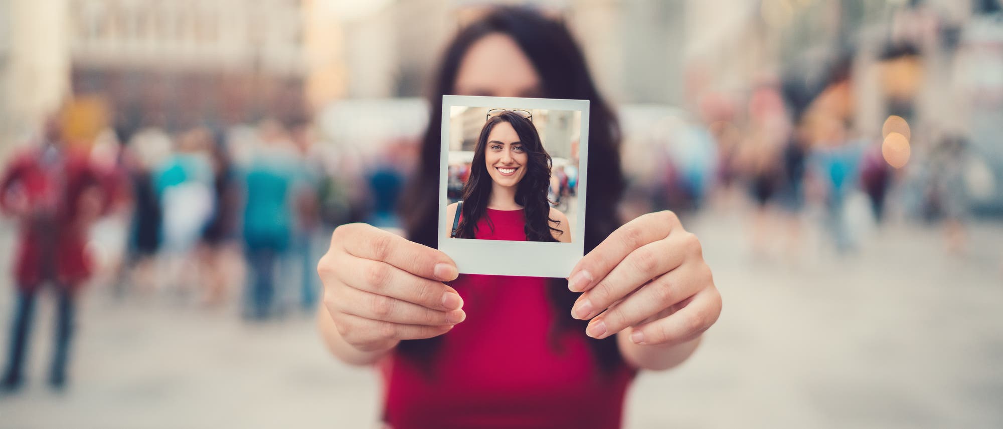 Eine Person in einem roten Kleid hält ein Polaroid-Foto vor sich, das ihr lächelndes Gesicht zeigt. Der Hintergrund ist eine belebte städtische Szene, die unscharf ist, um den Fokus auf das Foto zu lenken. Das Bild vermittelt ein Gefühl von Freude und urbanem Leben.
