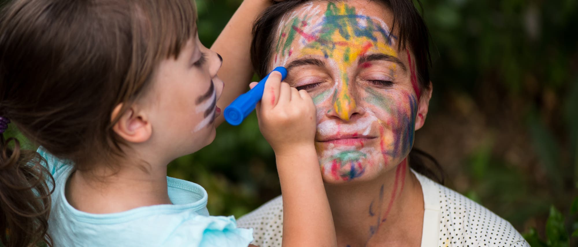 Ein Kind malt mit einem blauen Stift bunte Linien auf das Gesicht einer erwachsenen Person, die die Augen geschlossen h&auml;lt. Beide scheinen Spa&szlig; zu haben, w&auml;hrend sie im Freien sind. Die Szene vermittelt ein Gef&uuml;hl von Kreativit&auml;t und spielerischer Interaktion.