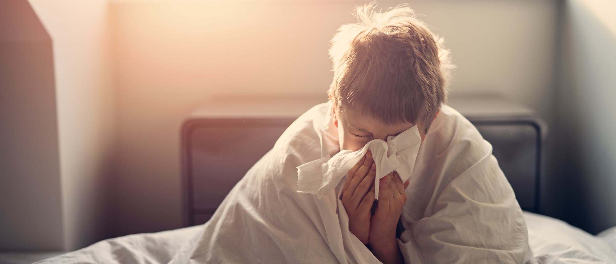 Ein Kind sitzt in einem Bett, eingehüllt in eine weiße Decke, und hält ein Taschentuch vor das Gesicht, als ob es niest. Sonnenlicht strömt durch ein Fenster im Hintergrund und erzeugt eine warme Atmosphäre.
