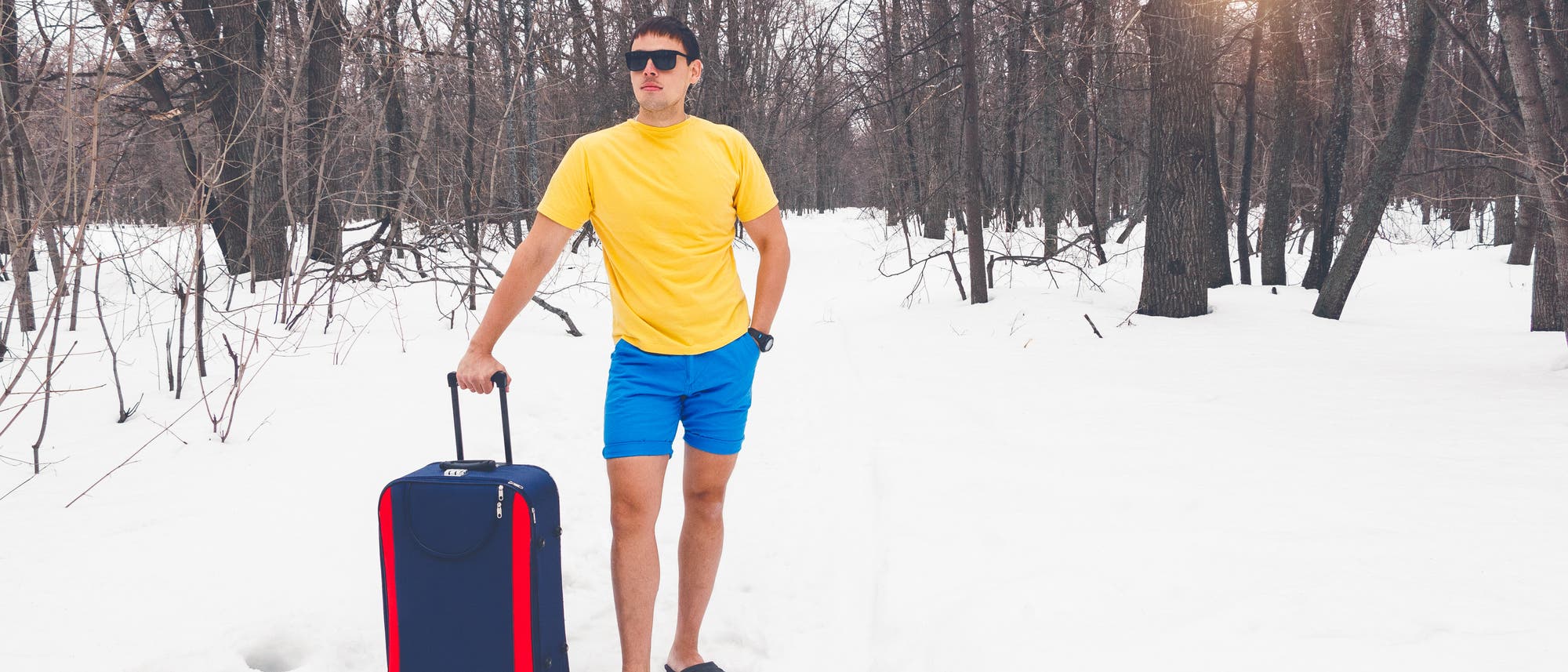 Ein Mann mit Sonnenbrille, gelbem T-Shirt, blauen Shorts und Sandalen steht in einem verschneiten Wald. Er h&auml;lt einen blauen Koffer mit zwei roten Streifen am Rand. Die B&auml;ume sind kahl, und die Sonne scheint durch die &Auml;ste. Der Kontrast zwischen der Sommerkleidung und der winterlichen Umgebung ist auff&auml;llig.