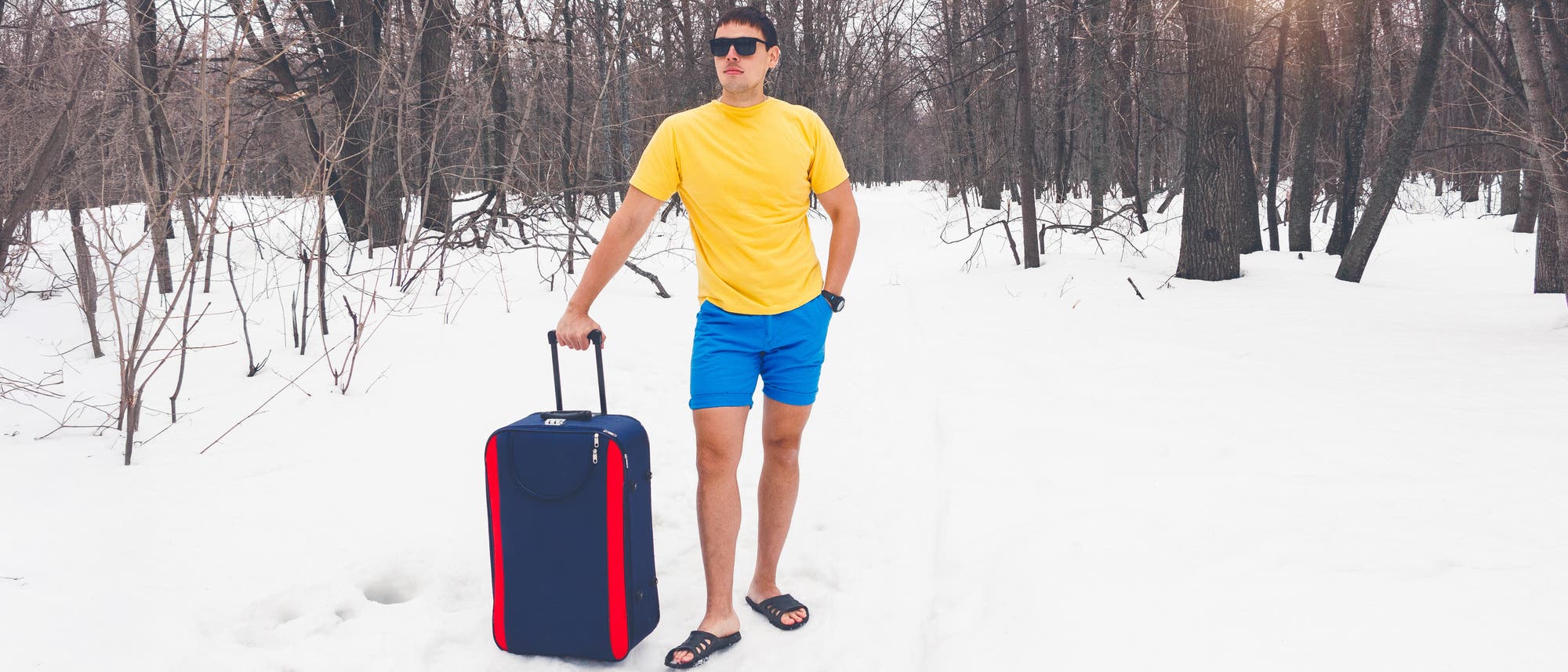 Ein Mann mit Sonnenbrille, gelbem T-Shirt, blauen Shorts und Sandalen steht in einem verschneiten Wald. Er hält einen blauen Koffer mit zwei roten Streifen am Rand. Die Bäume sind kahl, und die Sonne scheint durch die Äste. Der Kontrast zwischen der Sommerkleidung und der winterlichen Umgebung ist auffällig.