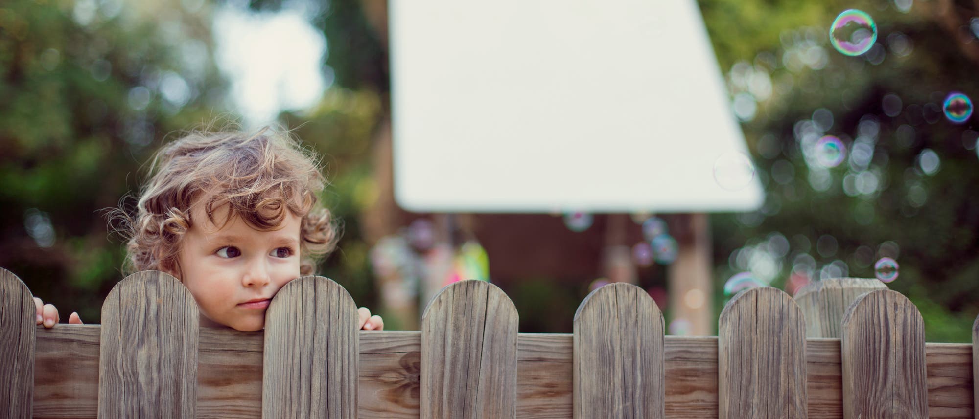 Ein Kind schaut neugierig &uuml;ber einen Holzzaun. Im Hintergrund sind unscharfe B&auml;ume und ein wei&szlig;es Schild zu sehen. Einige Seifenblasen schweben in der Luft. Die Szene vermittelt eine spielerische und neugierige Stimmung.
