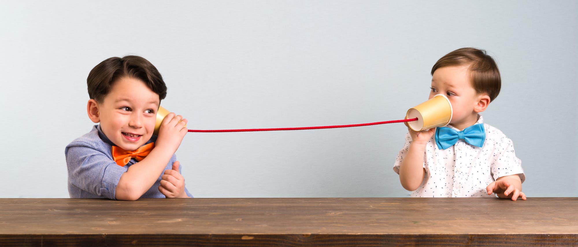 Zwei Kinder spielen mit einem selbst gemachten Telefon aus Pappbechern und einer Schnur. Das linke Kind hält einen Becher ans Ohr und lächelt, während das rechte Kind in den anderen Becher spricht. Beide tragen bunte Fliegen. Sie sitzen an einem Holztisch vor einem neutralen Hintergrund.
