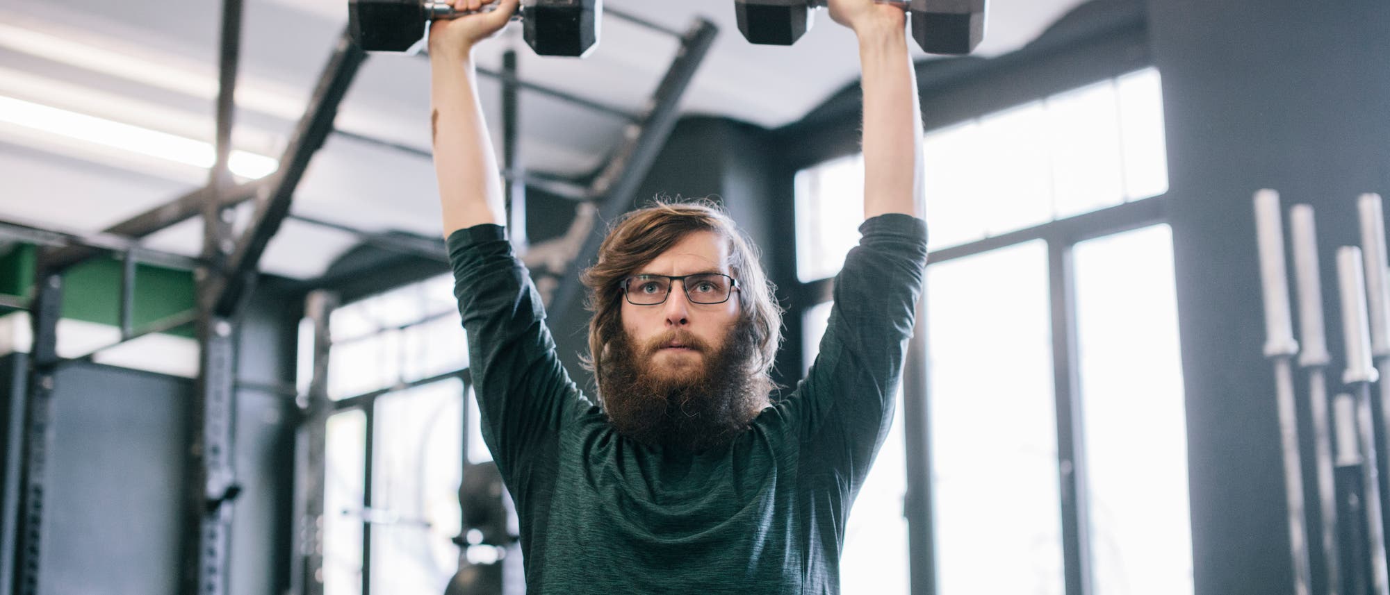 Ein Mann mit Brille und Bart hebt zwei Kurzhanteln über den Kopf in einem Fitnessstudio. Im Hintergrund sind Fitnessgeräte und große Fenster zu sehen. Die Szene vermittelt eine konzentrierte Trainingsatmosphäre. Ein Mann mit Brille und Bart hebt zwei Kurzhanteln über den Kopf in einem Fitnessstudio. Im Hintergrund sind Fitnessgeräte und große Fenster zu sehen. Die Szene vermittelt eine konzentrierte Trainingsatmosphäre.