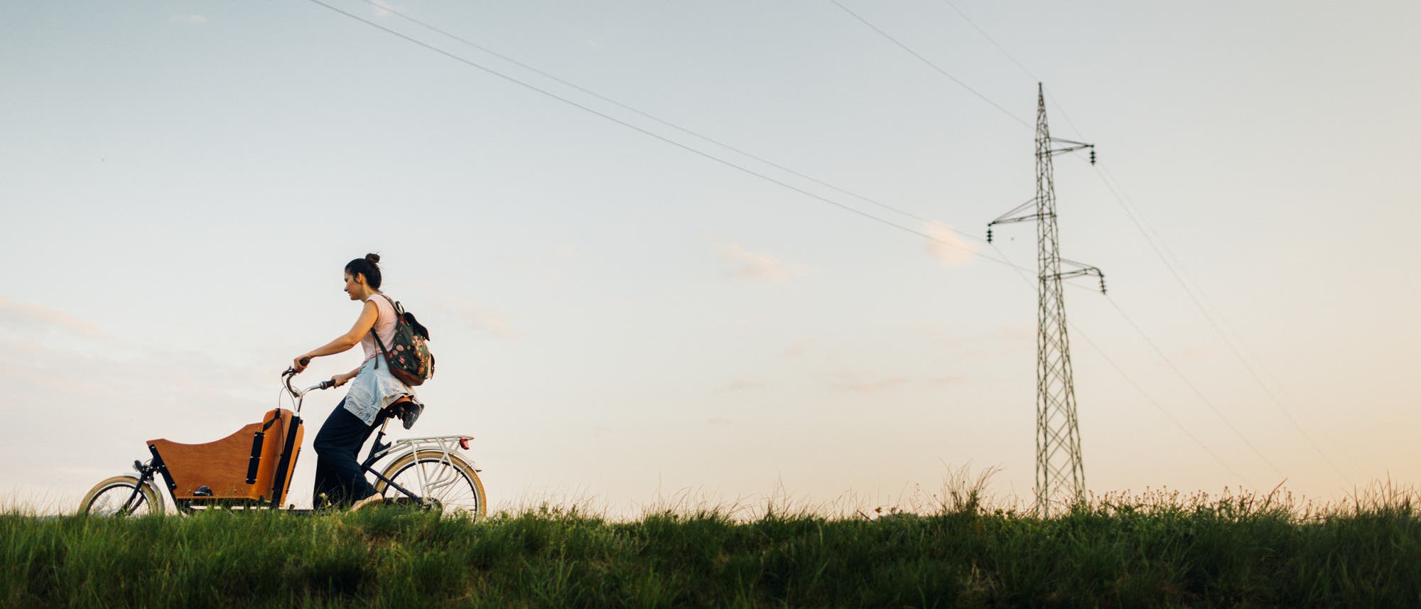 Eine Person f&auml;hrt bei Sonnenuntergang auf einem Lastenfahrrad einen grasbewachsenen H&uuml;gel entlang. Im Hintergrund ist ein Strommast zu sehen, der sich gegen den klaren Himmel abhebt. Die Szene vermittelt ein Gef&uuml;hl von Ruhe und Freiheit.