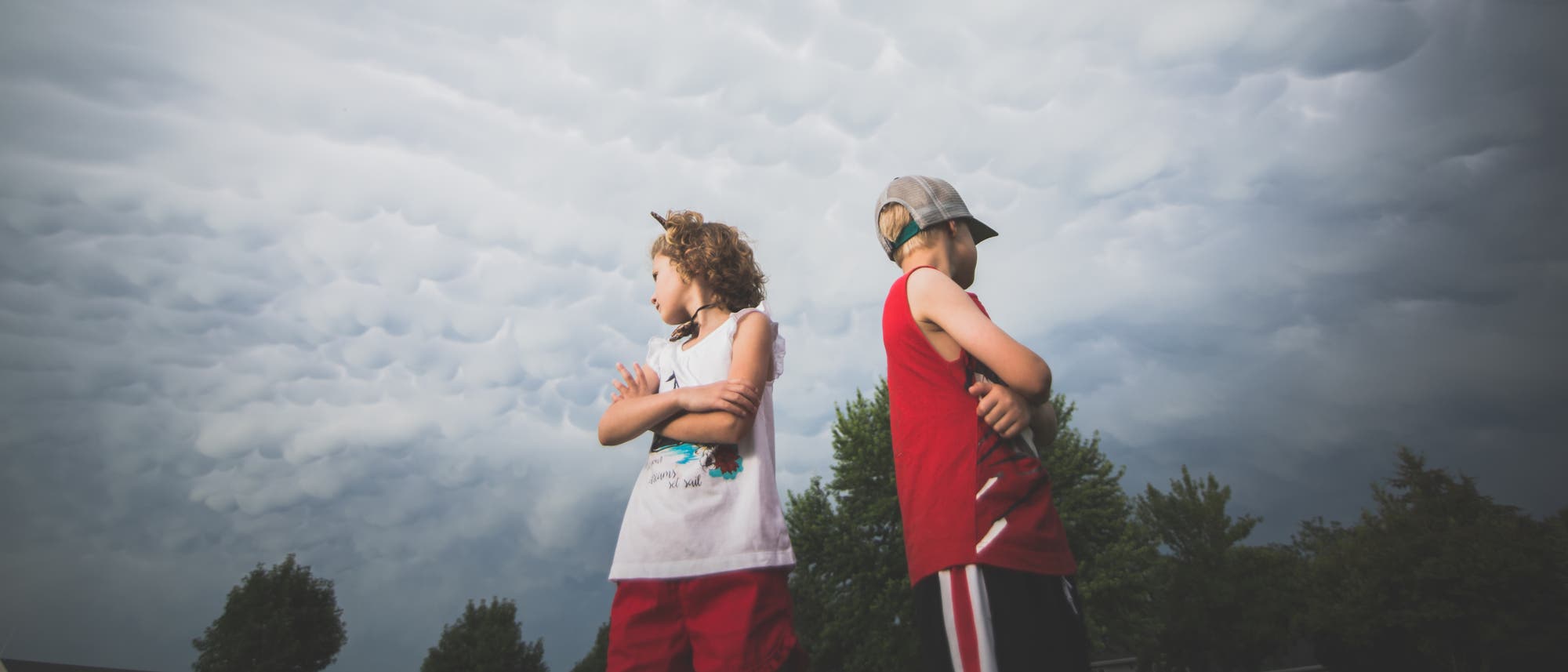 Zwei Kinder stehen Rücken an Rücken mit verschränkten Armen unter einem dramatischen Himmel mit Mammatuswolken. Beide schauen in entgegengesetzte Richtungen. Im Hintergrund sind Bäume zu sehen. Die Szene vermittelt eine nachdenkliche oder nachtragende Stimmung. Zwei Kinder stehen Rücken an Rücken mit verschränkten Armen unter einem dramatischen Himmel mit Mammatuswolken. Beide schauen in entgegengesetzte Richtungen. Im Hintergrund sind Bäume zu sehen. Die Szene vermittelt eine nachdenkliche oder nachtragende Stimmung.