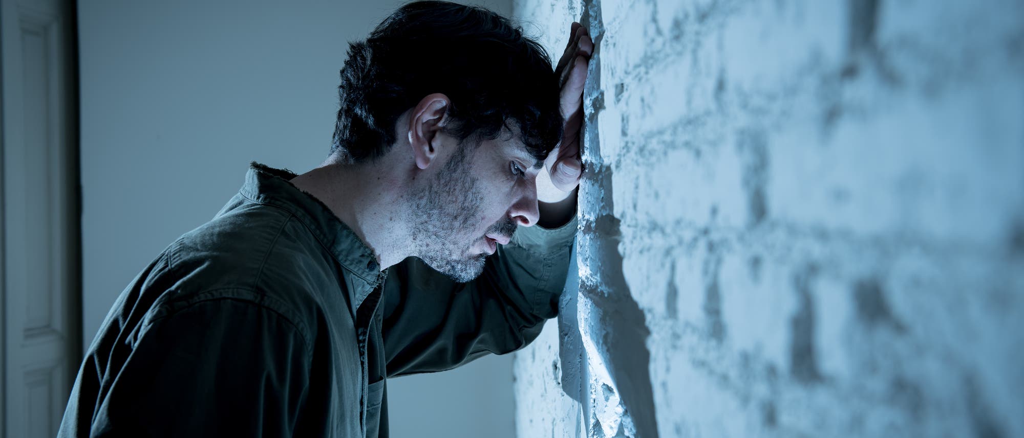 Ein Mann lehnt mit dem Kopf gegen eine weiße Backsteinwand, wirkt nachdenklich und bedrückt. Die Szene ist in kühlen Blautönen gehalten, was eine melancholische Stimmung vermittelt. Ein Mann lehnt mit dem Kopf gegen eine weiße Backsteinwand, wirkt nachdenklich und bedrückt. Die Szene ist in kühlen Blautönen gehalten, was eine melancholische Stimmung vermittelt.