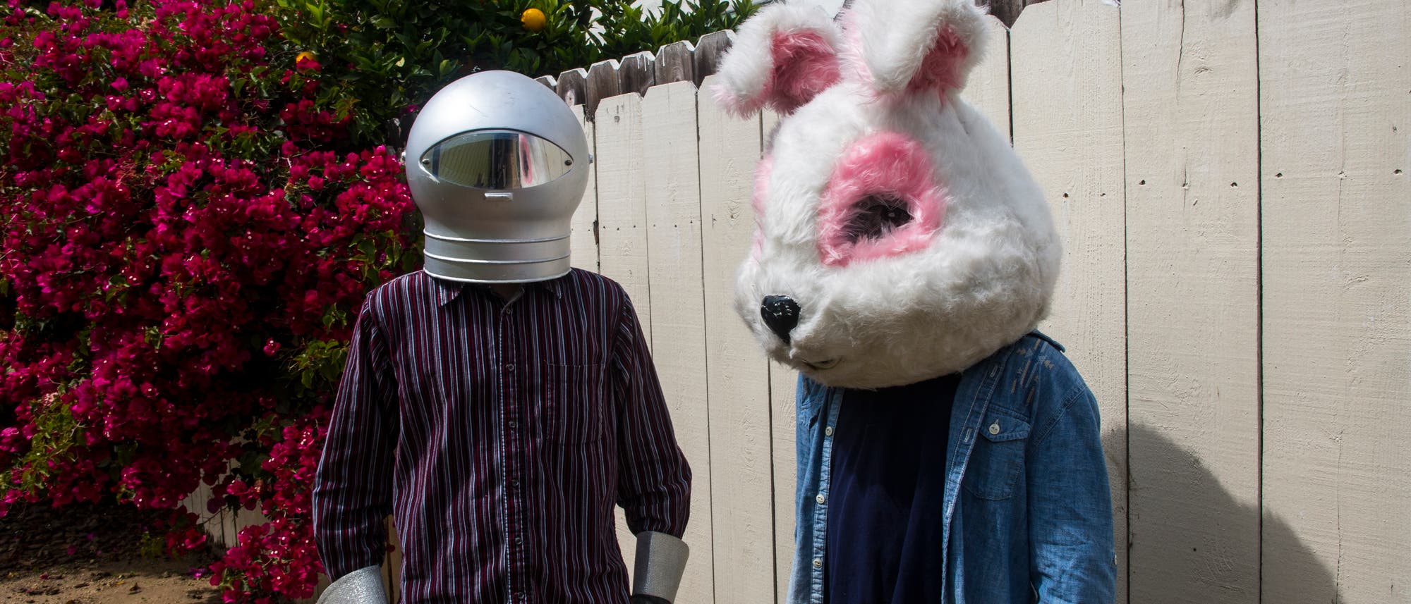 Zwei Personen stehen nebeneinander in einem Garten vor einem Holzzaun. Die linke Person tr&auml;gt einen silbernen Helm, der an einen Raumanzug erinnert, und eine gestreifte Bluse. Die rechte Person tr&auml;gt einen gro&szlig;en Hasenkopf mit rosa Details und eine Jeansjacke. Im Hintergrund sind bl&uuml;hende B&uuml;sche und ein wei&szlig;es Geb&auml;ude zu sehen.