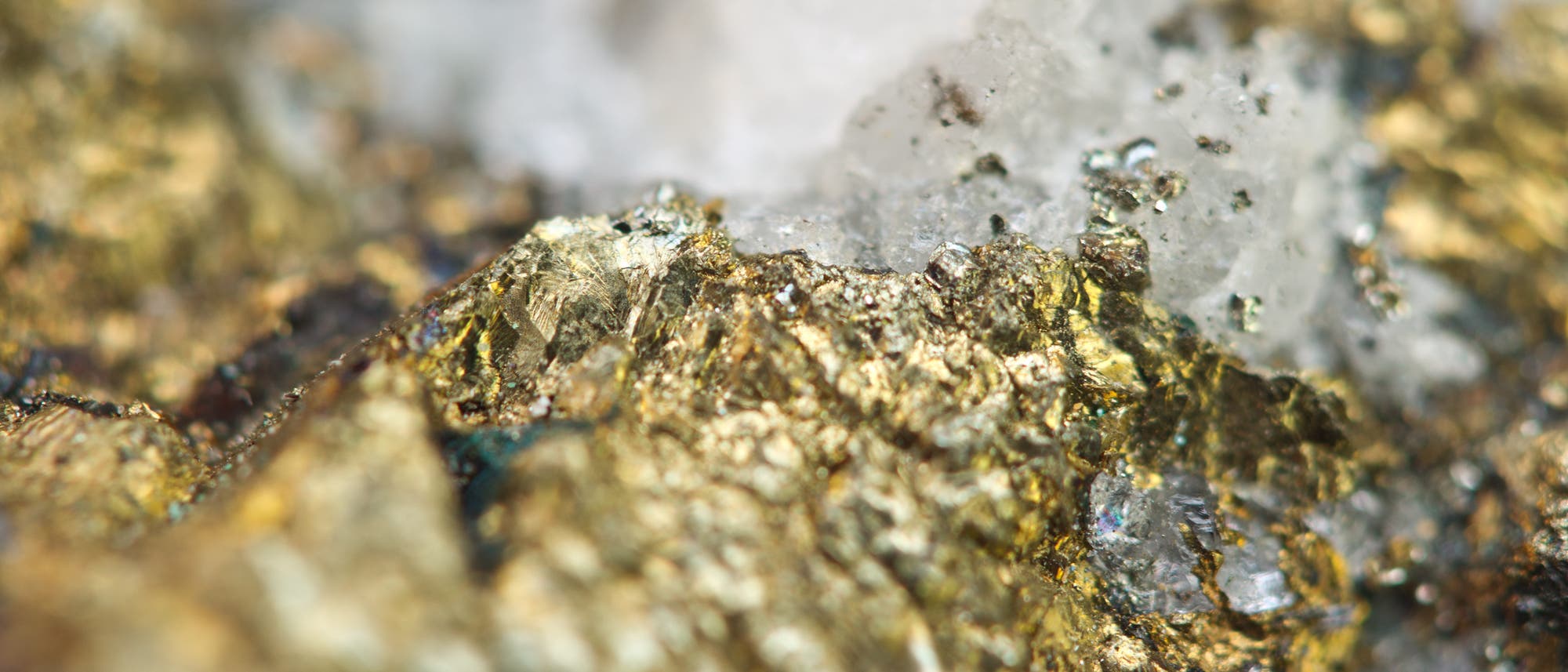 Nahaufnahme einer Gesteinsoberfl&auml;che mit goldenen und silbernen metallischen Reflexionen. Die Textur des Gesteins ist rau und unregelm&auml;&szlig;ig, mit gl&auml;nzenden, kristallinen Strukturen, die das Licht reflektieren. Im Hintergrund sind unscharfe Bereiche zu sehen, die die Tiefe der Oberfl&auml;che betonen.