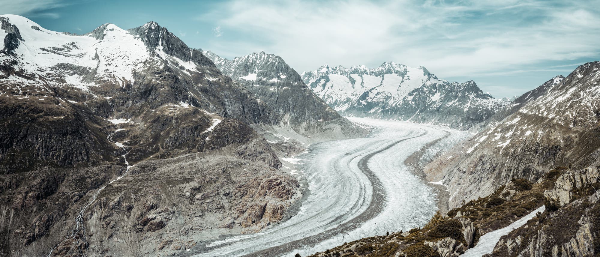 Eine weite Landschaftsaufnahme eines Gletschers, der sich durch ein Tal zwischen schneebedeckten Bergen schlängelt. Der Himmel ist leicht bewölkt, und die Berge zeigen felsige und schneebedeckte Flächen. Der Gletscher erstreckt sich in die Ferne und bildet eine markante, helle Linie in der Mitte des Bildes. Die Szene vermittelt eine ruhige, majestätische Atmosphäre. Eine weite Landschaftsaufnahme eines Gletschers, der sich durch ein Tal zwischen schneebedeckten Bergen schlängelt. Der Himmel ist leicht bewölkt, und die Berge zeigen felsige und schneebedeckte Flächen. Der Gletscher erstreckt sich in die Ferne und bildet eine markante, helle Linie in der Mitte des Bildes. Die Szene vermittelt eine ruhige, majestätische Atmosphäre.