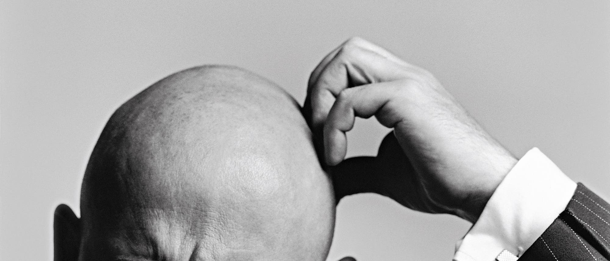 Ein Schwarz-Wei&szlig;-Foto zeigt eine Person, die sich nachdenklich den Kopf kratzt. Der Fokus liegt auf der Handbewegung und dem Ausdruck der Konzentration. Die Person tr&auml;gt einen Anzug mit einem gestreiften Muster. Der Hintergrund ist neutral und lenkt nicht vom Hauptmotiv ab.