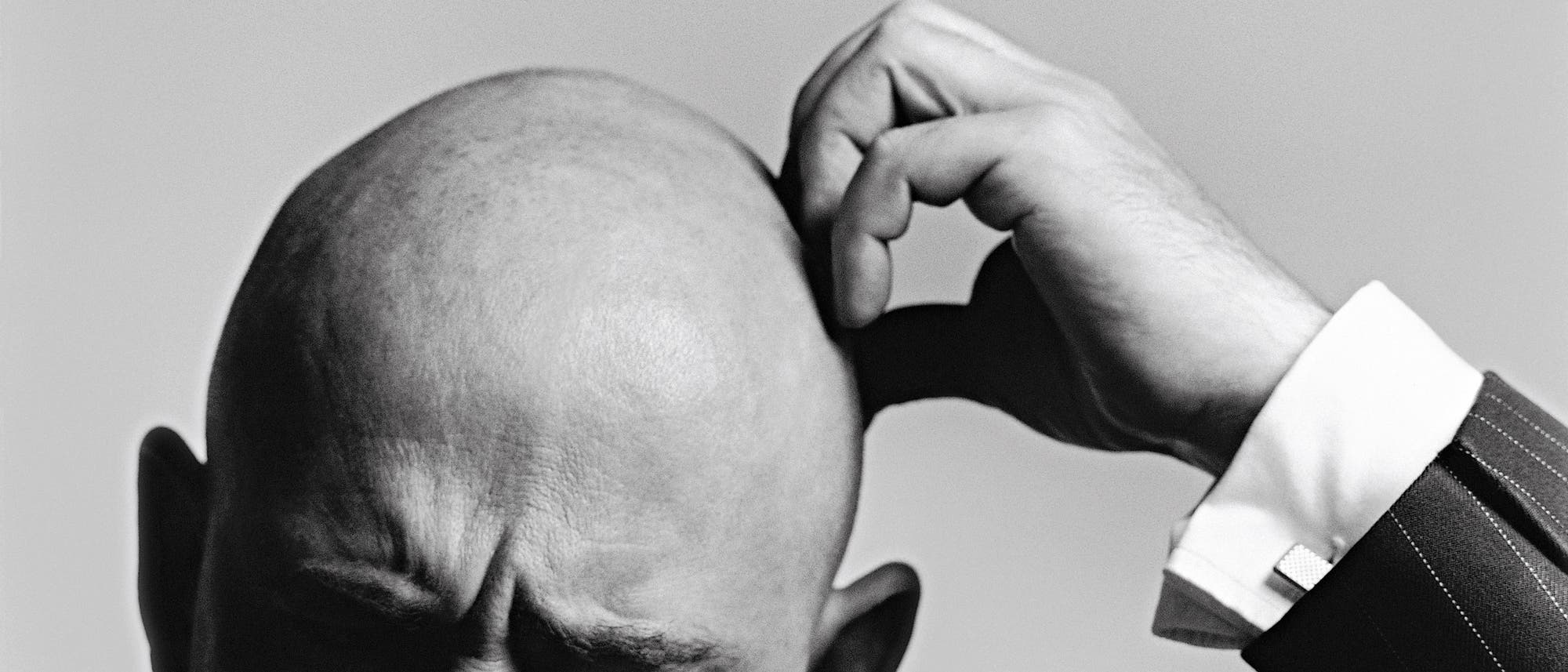 Ein Schwarz-Wei&szlig;-Foto zeigt eine Person, die sich nachdenklich den Kopf kratzt. Der Fokus liegt auf der Handbewegung und dem Ausdruck der Konzentration. Die Person tr&auml;gt einen Anzug mit einem gestreiften Muster. Der Hintergrund ist neutral und lenkt nicht vom Hauptmotiv ab.