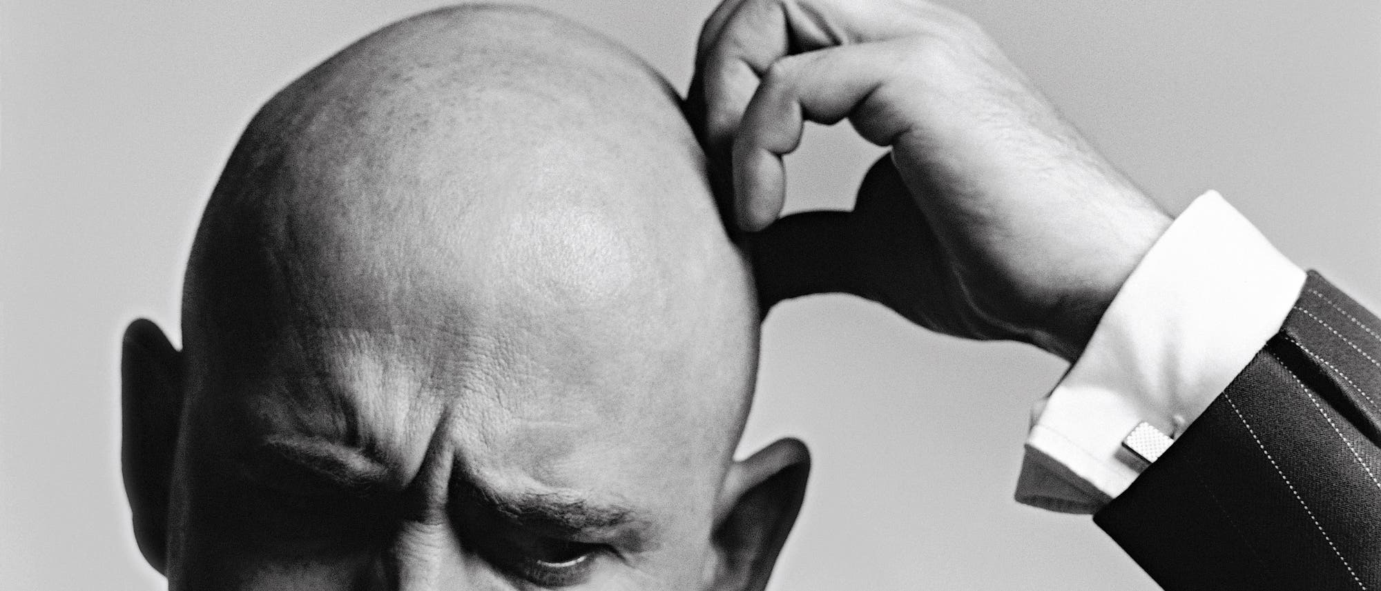 Ein Schwarz-Weiß-Foto zeigt eine Person, die sich nachdenklich den Kopf kratzt. Der Fokus liegt auf der Handbewegung und dem Ausdruck der Konzentration. Die Person trägt einen Anzug mit einem gestreiften Muster. Der Hintergrund ist neutral und lenkt nicht vom Hauptmotiv ab.