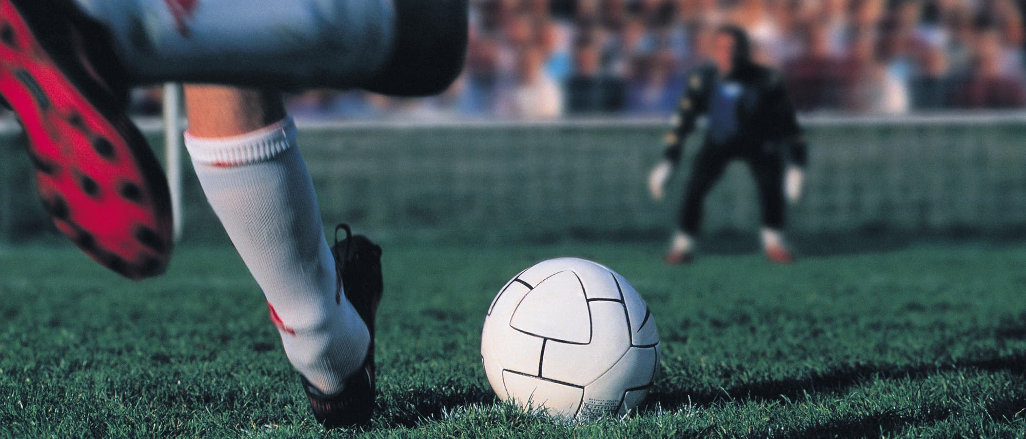 Ein Fußballspieler in weißen Stutzen und roten Schuhen tritt auf einem grünen Spielfeld gegen einen Fußball. Im Hintergrund steht ein Torwart vor dem Tor, bereit, den Ball abzuwehren. Die Szene findet in einem Stadion mit unscharfem Publikum im Hintergrund statt. Ein Fußballspieler in weißen Stutzen und roten Schuhen tritt auf einem grünen Spielfeld gegen einen Fußball. Im Hintergrund steht ein Torwart vor dem Tor, bereit, den Ball abzuwehren. Die Szene findet in einem Stadion mit unscharfem Publikum im Hintergrund statt.