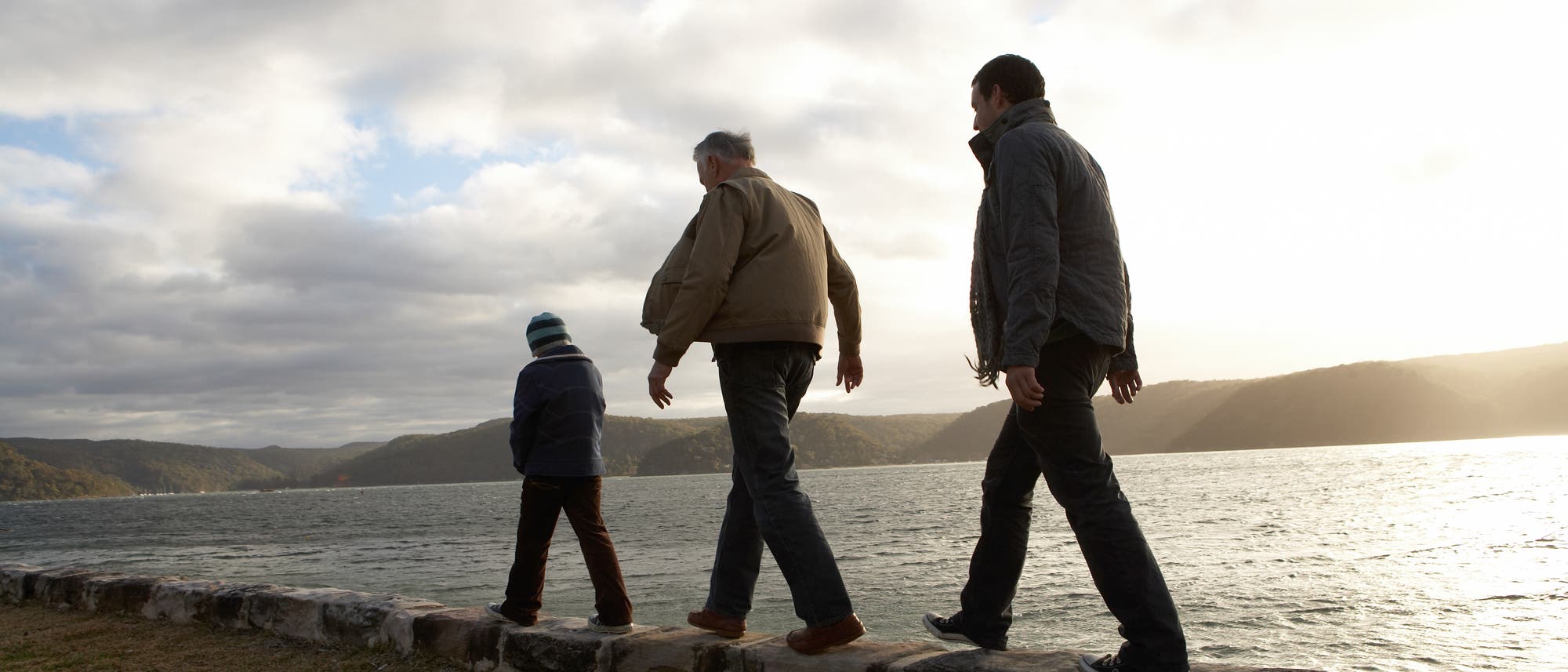 Drei Personen verschiedenen Alters – vermutlich Opa, Vater und Enkel – spazieren hintereinander auf einer niedrigen Steinmauer entlang eines Gewässers. Der Himmel ist bewölkt, und die Sonne scheint durch die Wolken. Im Hintergrund sind bewaldete Hügel zu sehen. Drei Personen verschiedenen Alters – vermutlich Opa, Vater und Enkel – spazieren hintereinander auf einer niedrigen Steinmauer entlang eines Gewässers. Der Himmel ist bewölkt, und die Sonne scheint durch die Wolken. Im Hintergrund sind bewaldete Hügel zu sehen.