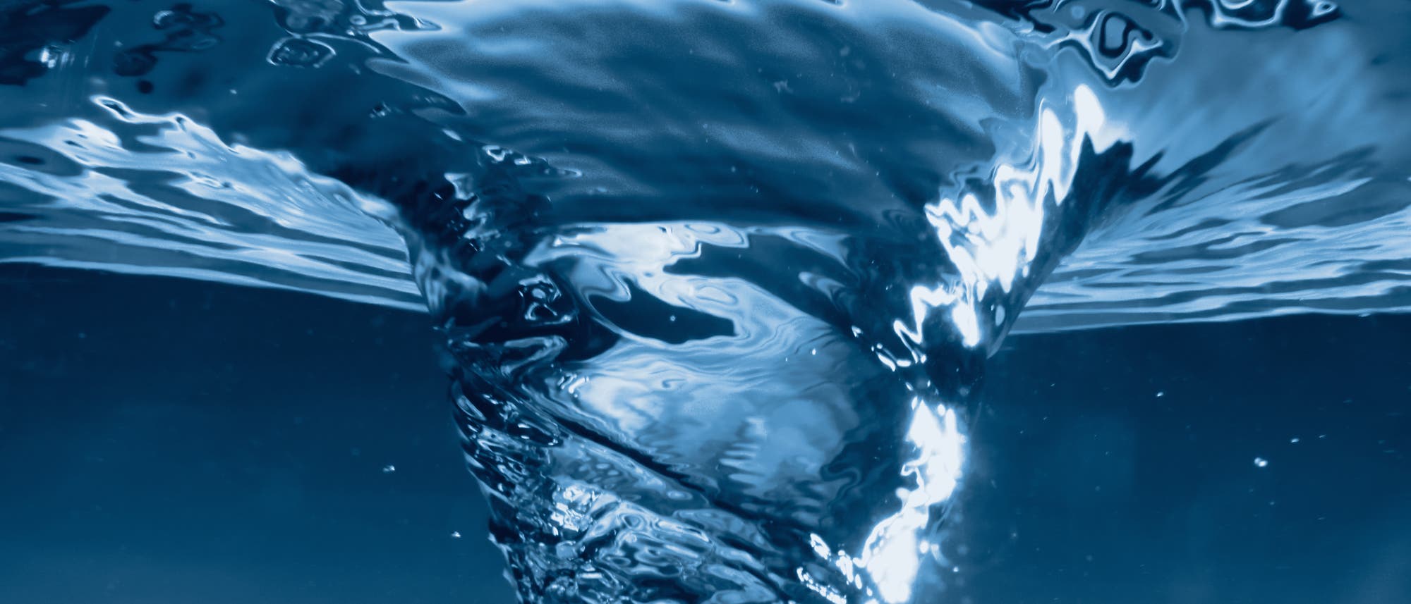 Ein Wirbel aus klarem, blauem Wasser, der sich spiralförmig nach unten bewegt. Die Lichtreflexionen auf der Wasseroberfläche erzeugen glänzende, wellenförmige Muster. 