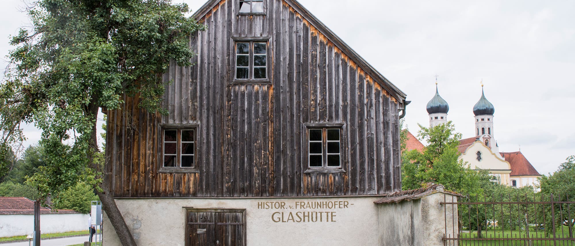 Ein altes, hölzernes Gebäude mit der Aufschrift „Histor. v. Fraunhofer Glashütte“ auf der Fassade. Links steht ein Baum, und im Hintergrund sind die Zwiebeltürme einer Kirche zu sehen. Der Himmel ist bewölkt, und das Gebäude steht auf einer grünen Wiese. Ein altes, hölzernes Gebäude mit der Aufschrift „Histor. v. Fraunhofer Glashütte“ auf der Fassade. Links steht ein Baum, und im Hintergrund sind die Zwiebeltürme einer Kirche zu sehen. Der Himmel ist bewölkt, und das Gebäude steht auf einer grünen Wiese.