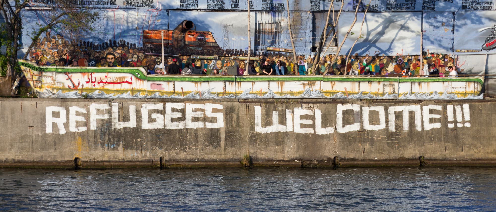 Auf eine Mauer am Rand eines Flusses befindet sich ein Graffito "Refugees Welcome".